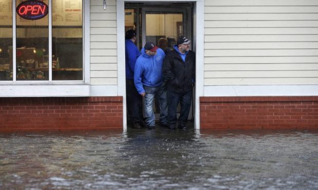 People stand at the entrance to a pizza shop as water floods a street, in Scituate, Mass., Friday, ...