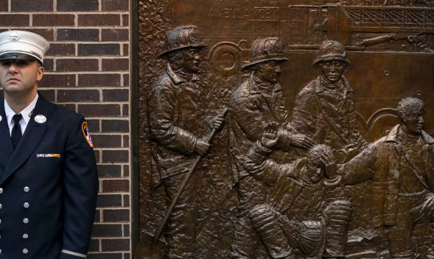 A New York City firefighter stands at attention in front of a memorial on the side of a firehouse a...